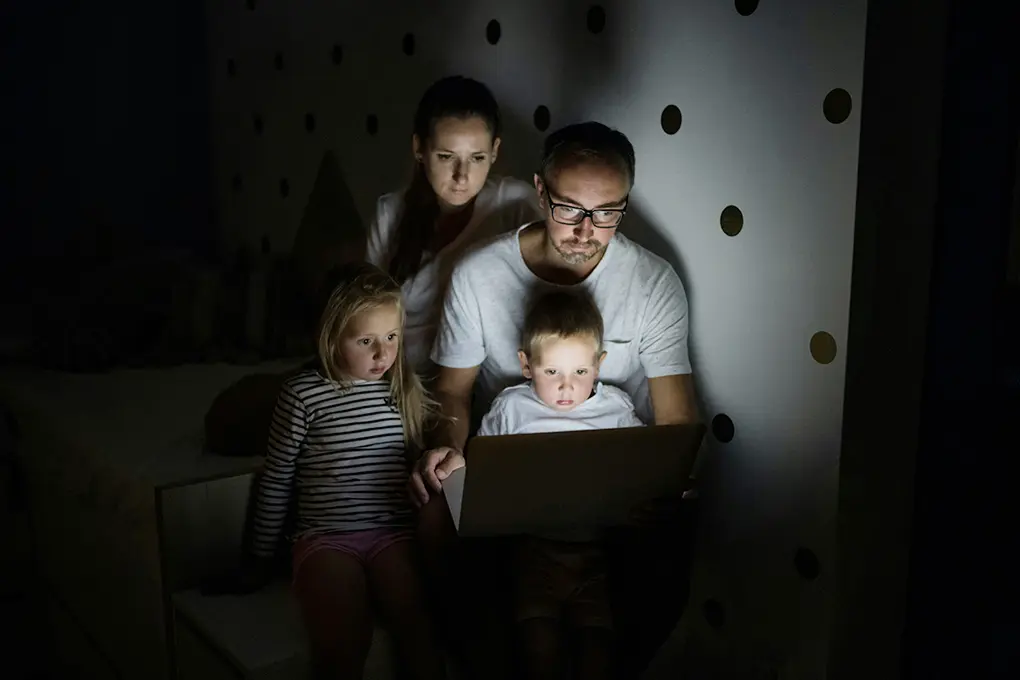 A couple and their two children sit in a dimly lit room, focused on a laptop screen displaying their smart home security system