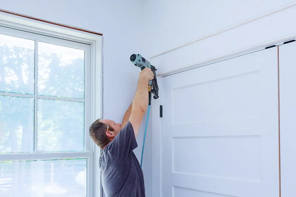 A person in a gray shirt uses a nail gun to install trim near a white door in a bright room