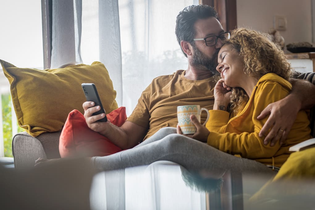 A couple sits on a couch, cuddling and smiling. The man holds a smartphone, while the woman rests her head on his shoulder, holding a mug