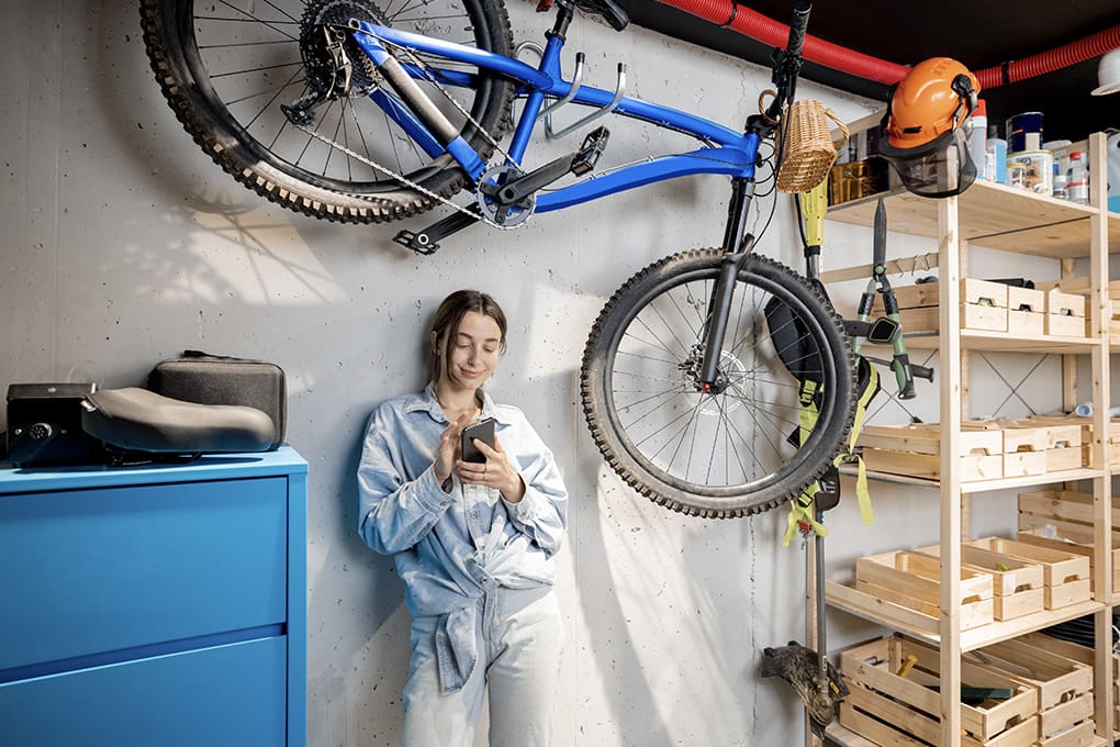 A woman leans against a wall, smiling at her phone, with a blue bike mounted above and some shelves and boxes in the garage