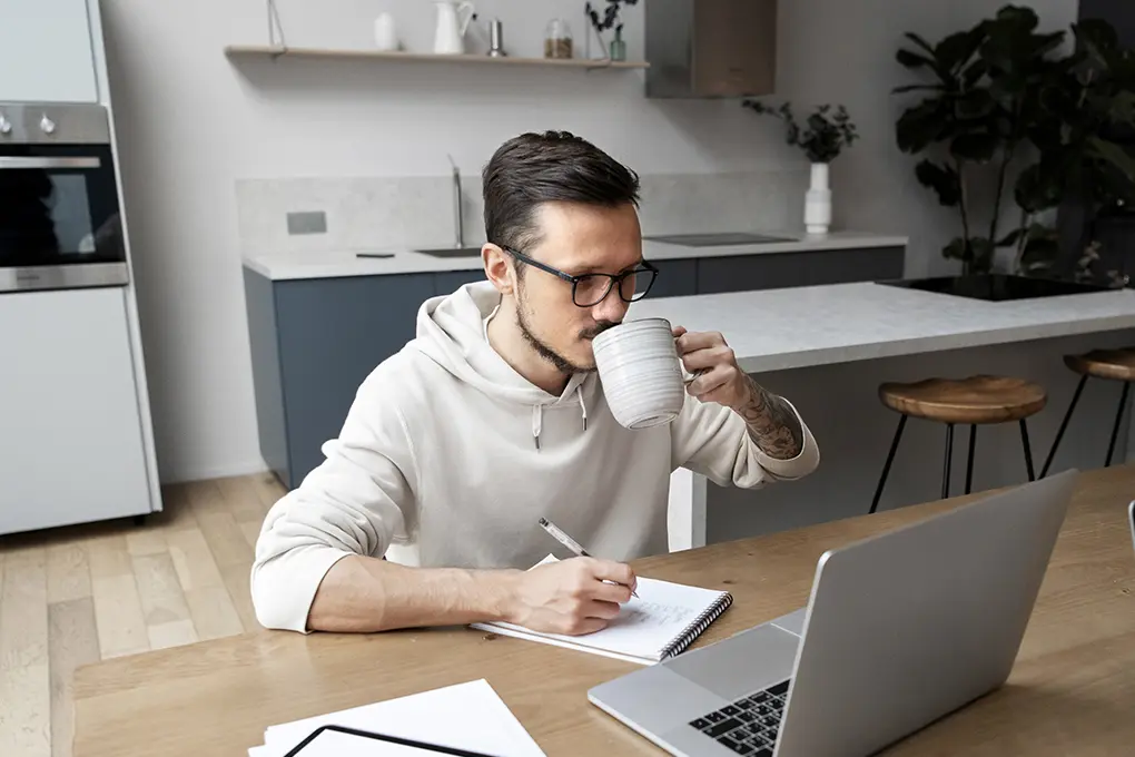A man in glasses sits at a wooden table, sipping coffee while jotting notes. A laptop is open, conveying a focused, cozy mood.